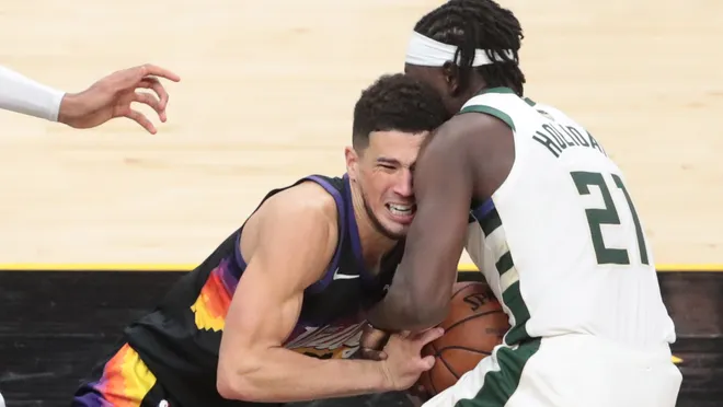 Jrue Holiday making a pivotal defensive play in Game 5 of the 2021 NBA Finals, stripping the ball from Devin Booker of the Phoenix Suns. Wearing a Milwaukee Bucks jersey, Holiday is in motion, delivering an alley-oop pass to Giannis Antetokounmpo that sealed the game and swung the series momentum. This defining moment led to the Bucks' first NBA championship in 50 years, a key highlight covered by Matt Goren for All Angles.